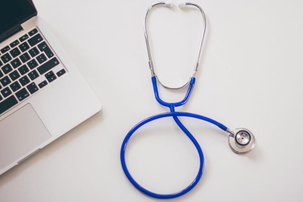Blue stethoscope beside silver laptop on white surface, symbolizing health tech.