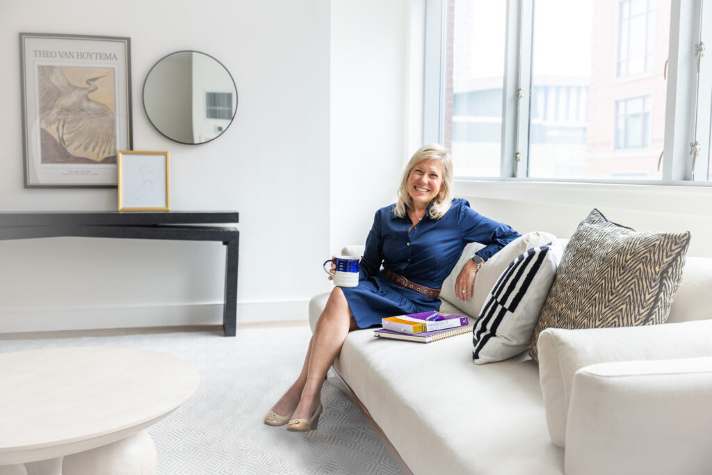 Dr. Jennifer Schwartz, a telehealth weight loss doctor, in blue dress on sofa, holding mug, with cushions and books.